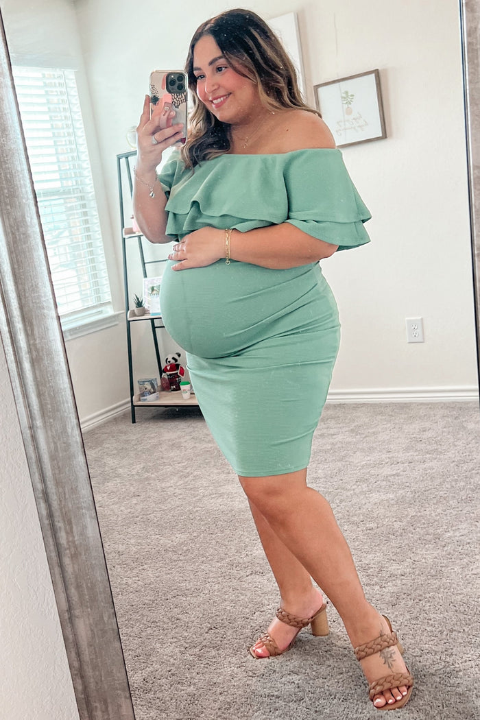 woman wearing a light green off-the-shoulder maternity dress, showing her baby bump while smiling in front of a mirror