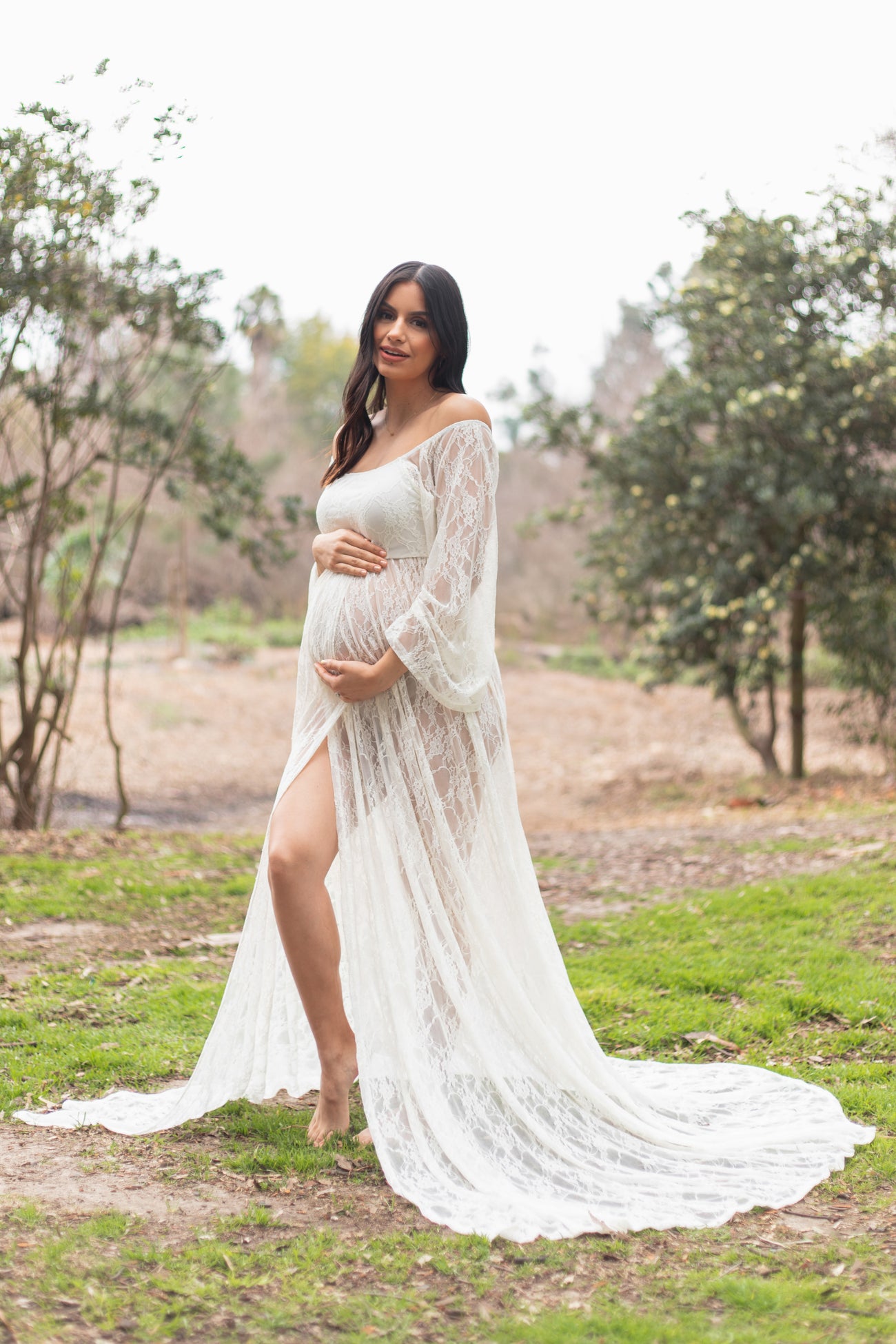 woman wearing a white lace maternity gown with a thigh-high slit, gently cradling her baby bump outdoors