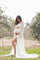 woman wearing a white lace maternity gown with flowing sleeves, holding her baby bump in a natural outdoor setting