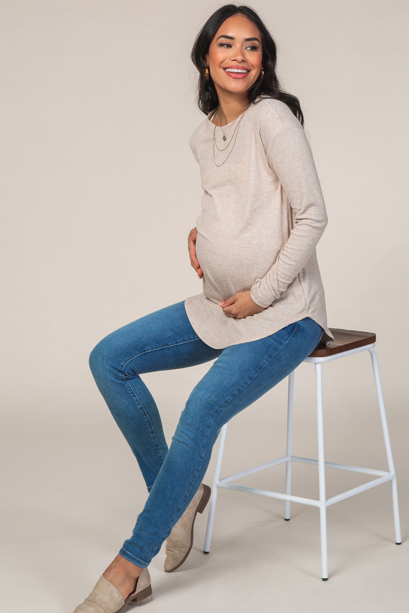 woman wearing a beige long sleeve maternity top and blue skinny jeans, showing her baby bump while sitting on a stool