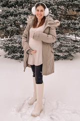 smiling pregnant woman in winter parka and earmuffs standing in snow by pine trees