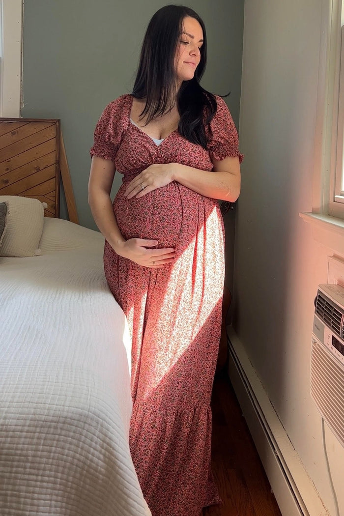 woman wearing a floral pink maternity dress cradling her baby bump while standing by a window