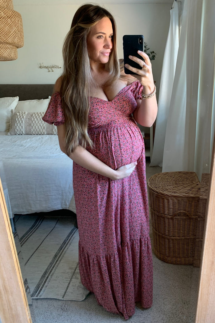 woman wearing a pink floral off-the-shoulder maternity dress and holding her baby bump