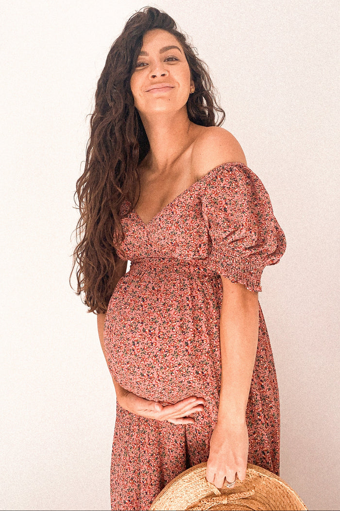 woman wearing a floral off-shoulder maternity dress, showcasing her baby bump and holding a straw hat