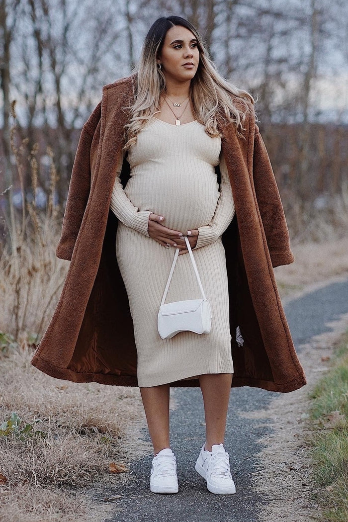 woman wearing a beige ribbed maternity dress and a brown oversized coat, holding her baby bump, styled with a white purse and sneakers