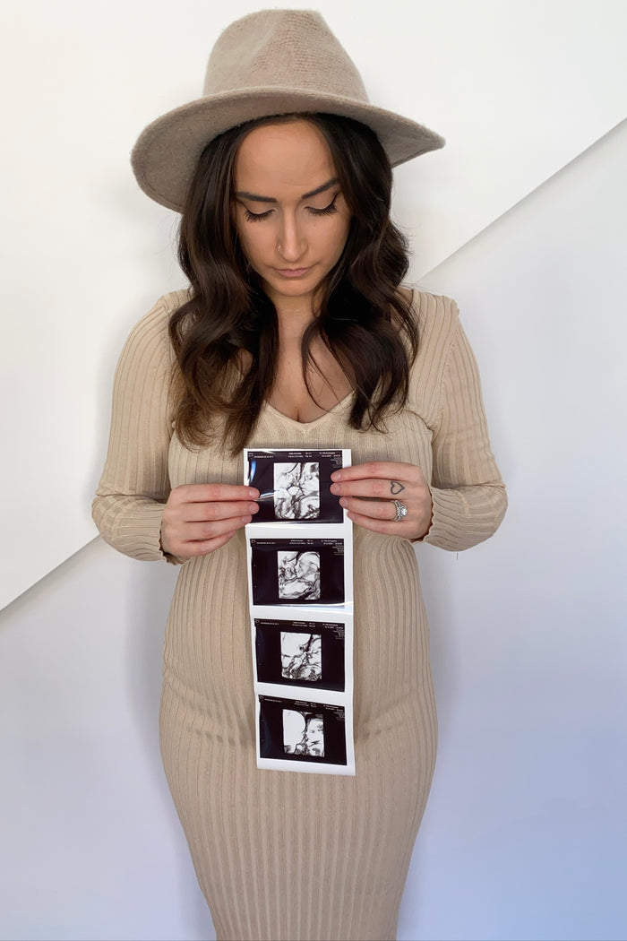 woman wearing a beige ribbed maternity dress and holding ultrasound photos while standing against a white background