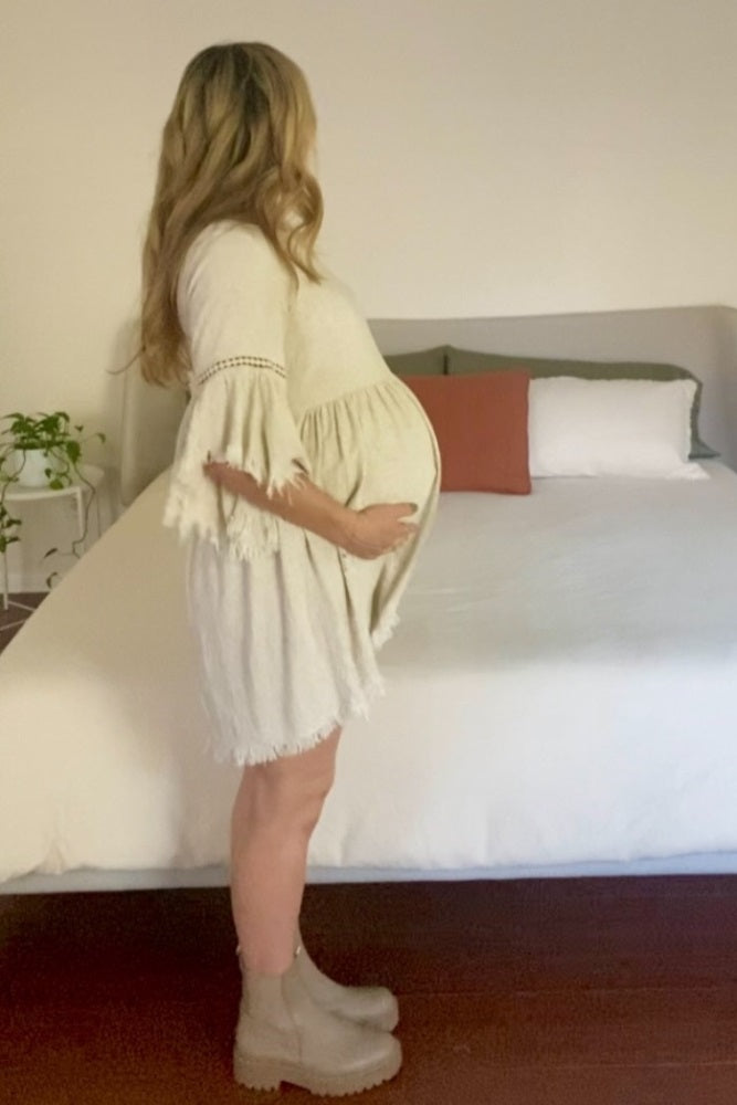 woman wearing a cream short sleeve maternity dress with fringe details and holding her baby bump while standing beside a bed