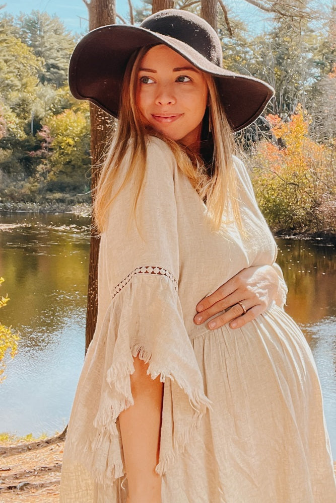 pregnant woman in a beige linen maternity dress and black sun hat posing by a river in autumn