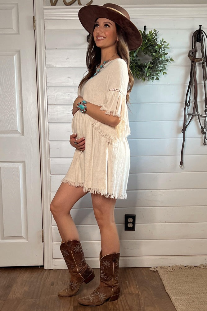 woman wearing a cream maternity dress with fringe details and cowboy boots, showcasing her baby bump