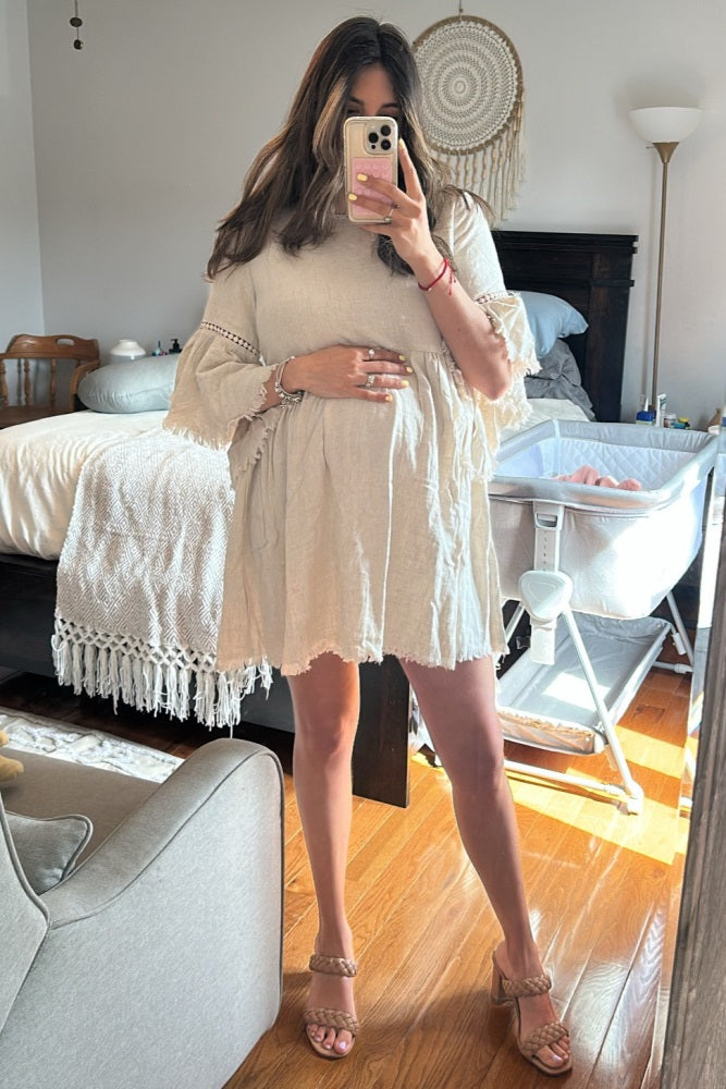 woman wearing a light beige maternity dress with fringe detail and holding her baby bump