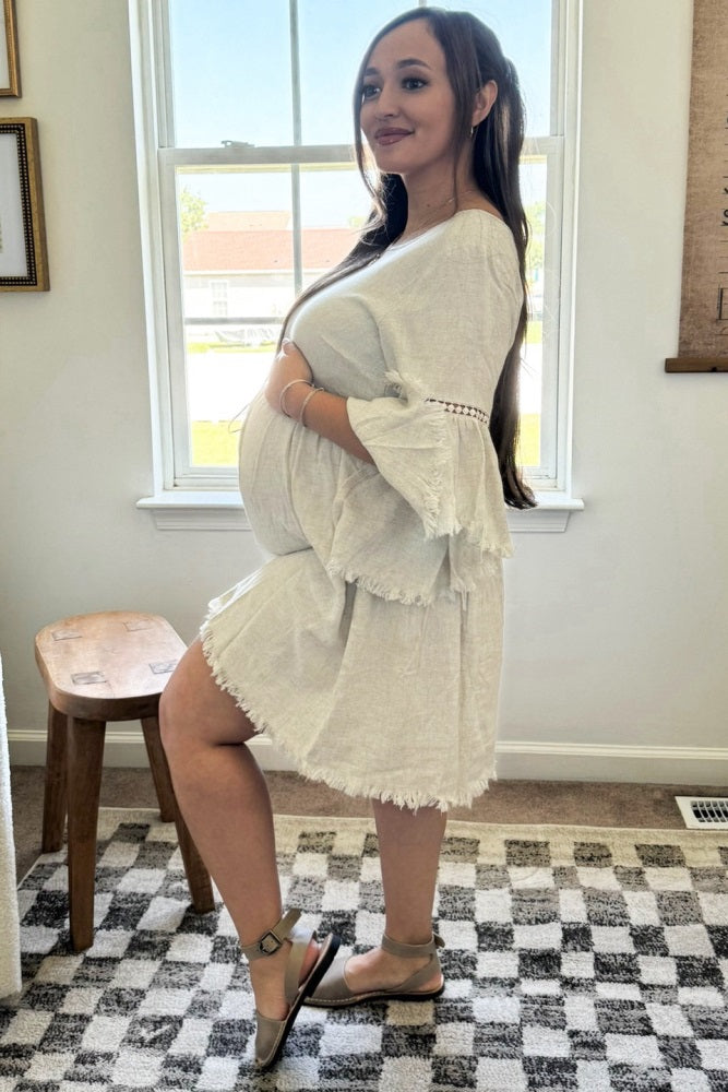 woman wearing a white maternity dress with bell sleeves and frayed hem, cradling her baby bump
