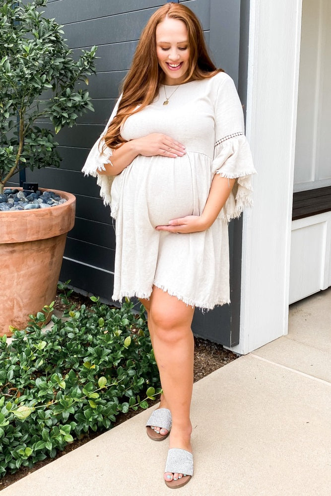 woman wearing a light beige short sleeve maternity dress and holding her baby bump outside.