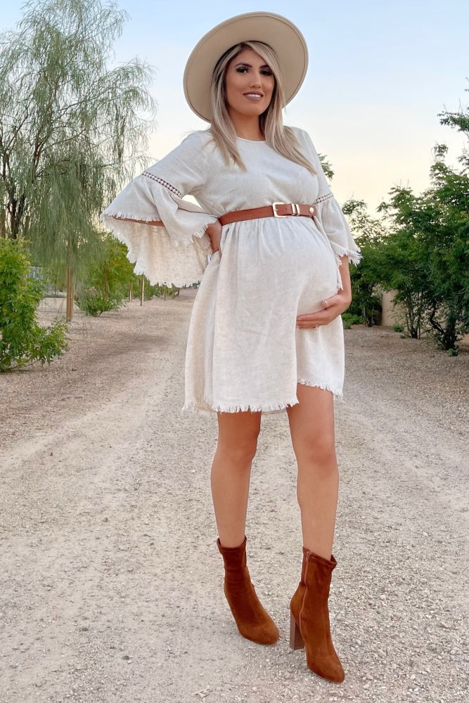 woman wearing a beige maternity dress with bell sleeves and a brown belt, holding her baby bump, paired with brown ankle boots and a hat