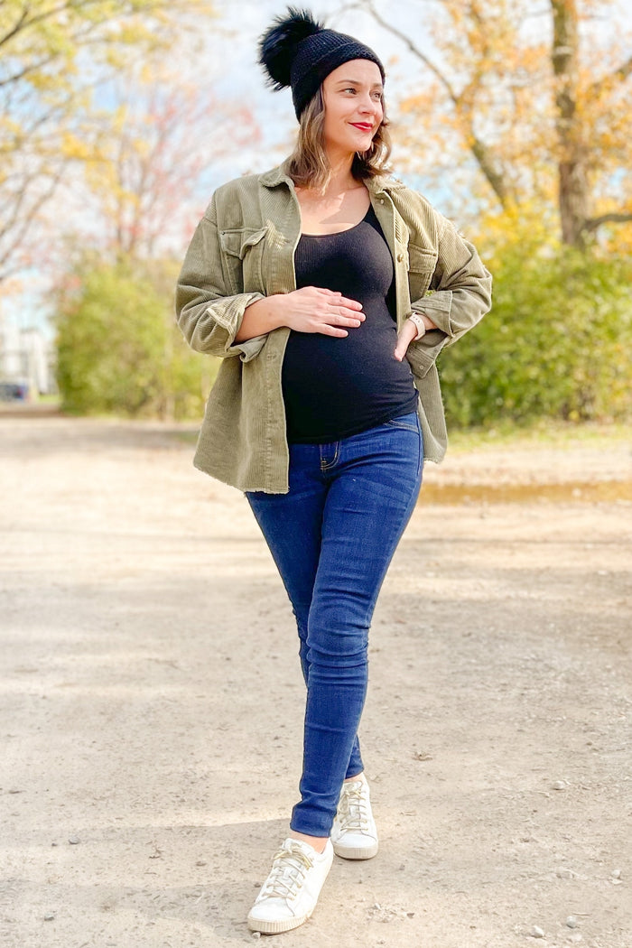 woman wearing a black maternity tank top, olive green jacket, and blue skinny jeans, showing her baby bump outdoors