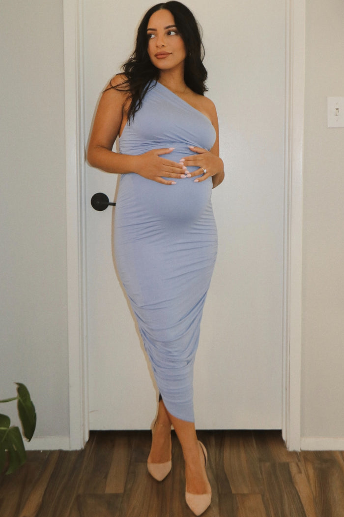 woman wearing a light blue one-shoulder maternity dress and holding her baby bump