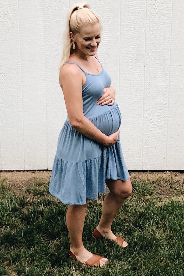 woman wearing a light blue maternity dress with a tiered skirt and holding her baby bump while standing on grass