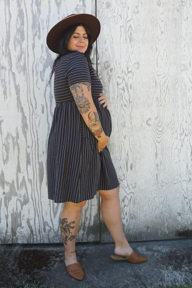woman wearing a black and white striped maternity dress and holding her baby bump, accessorized with a hat and sandals