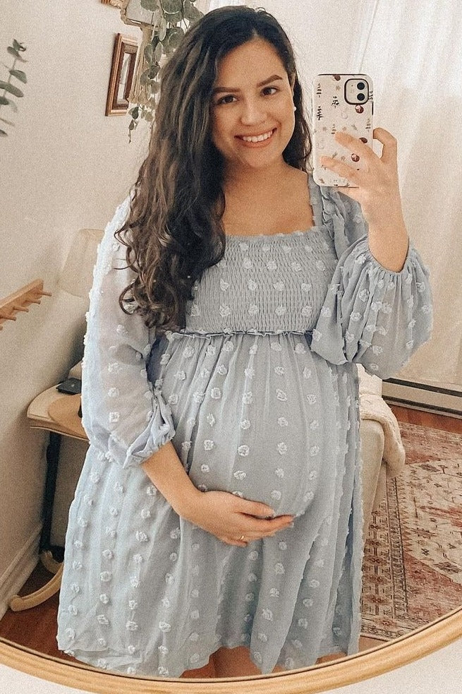 woman wearing a light blue maternity dress with puff sleeves, smiling and holding her baby bump in front of a mirror