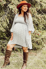 woman wearing a light gray maternity dress with puff sleeves and polka dot details, showing her baby bump, paired with a brown hat and tall boots