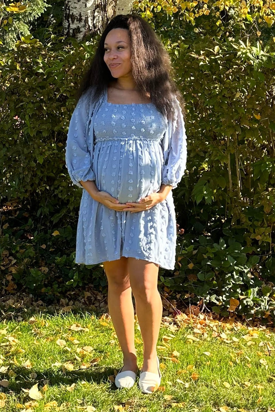 woman wearing a light blue maternity dress with puff sleeves and holding her baby bump in a garden setting