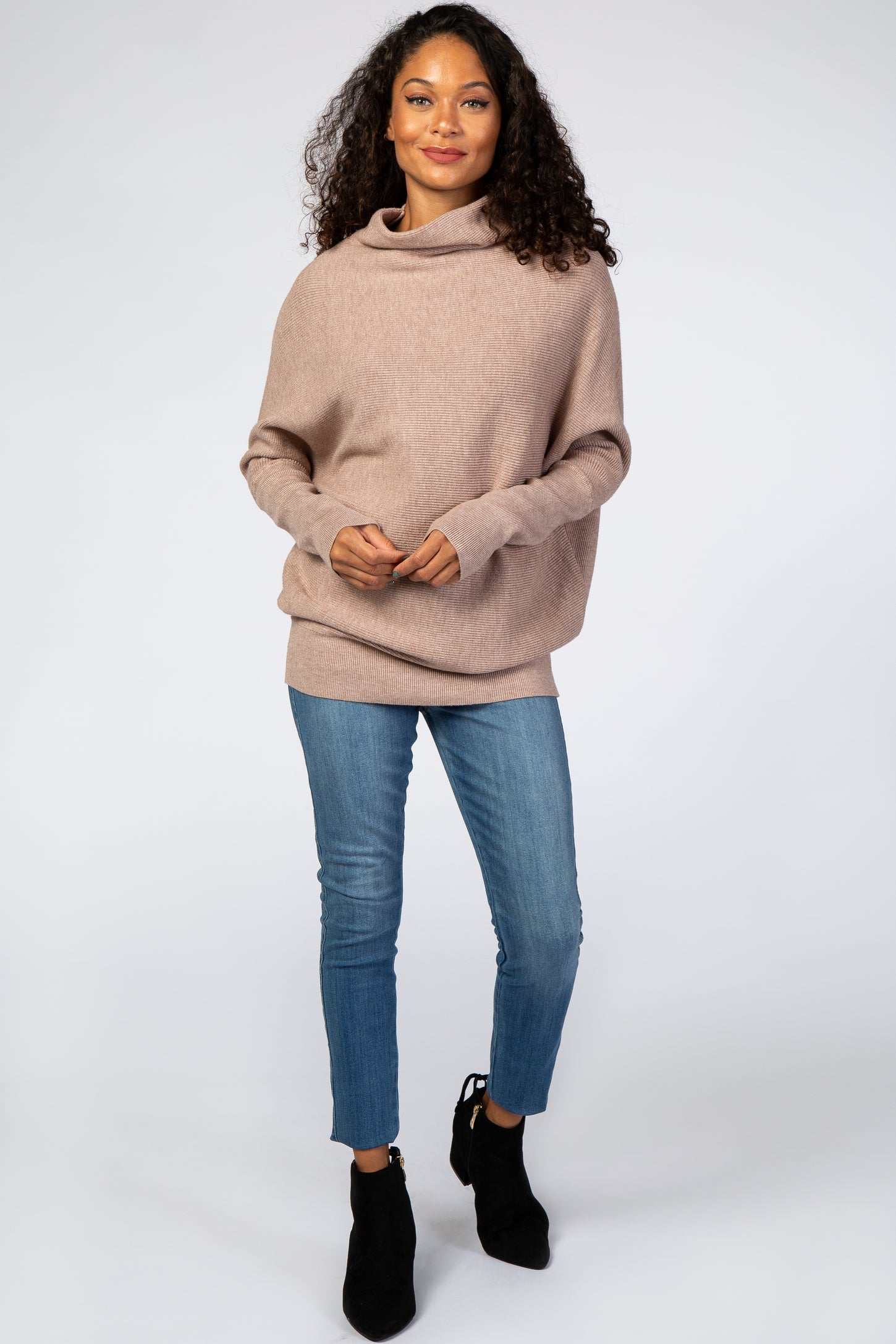 woman wearing a taupe oversized cowl neck sweater and blue skinny jeans with black ankle boots