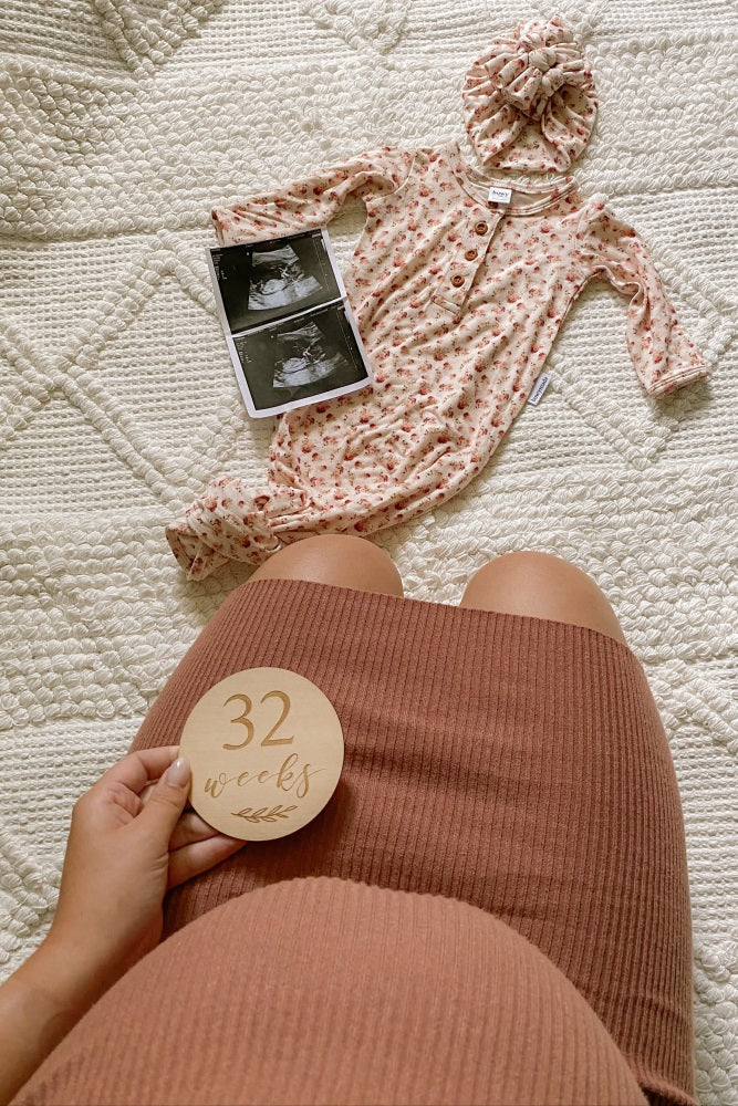 Pregnant woman holding 32 weeks milestone disc next to floral baby gown and ultrasound photos
