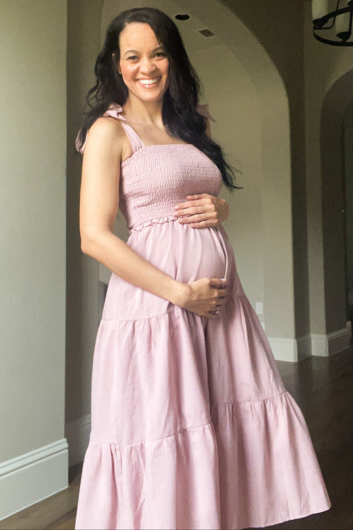 smiling pregnant woman in a pink smocked maternity dress holding her baby bump indoors