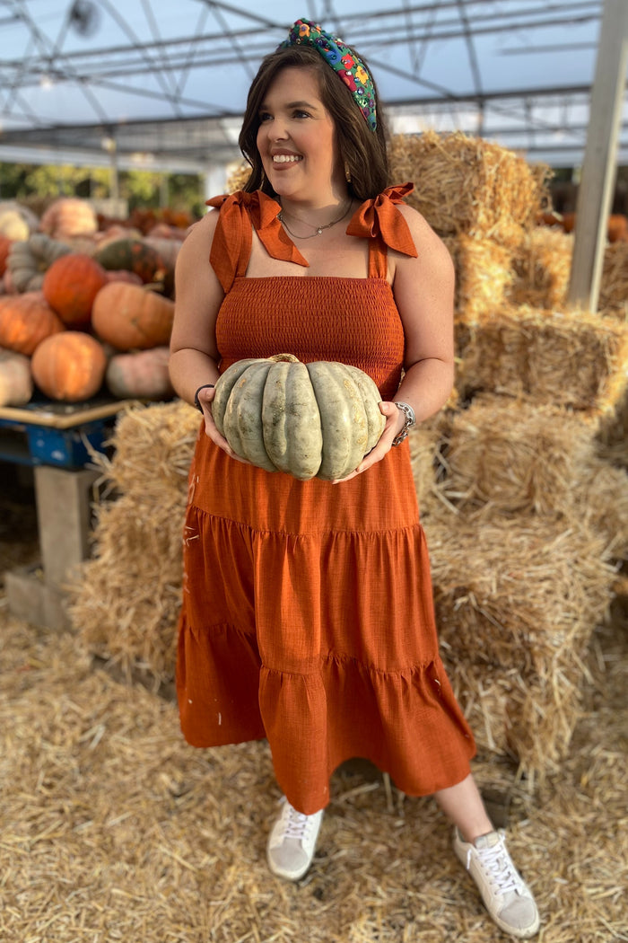 woman wearing an orange maternity dress with tie straps, holding a pumpkin in a pumpkin patch