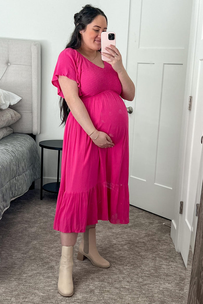 woman wearing a hot pink maternity dress with a flowy skirt and short sleeves, holding her baby bump in front of a mirror