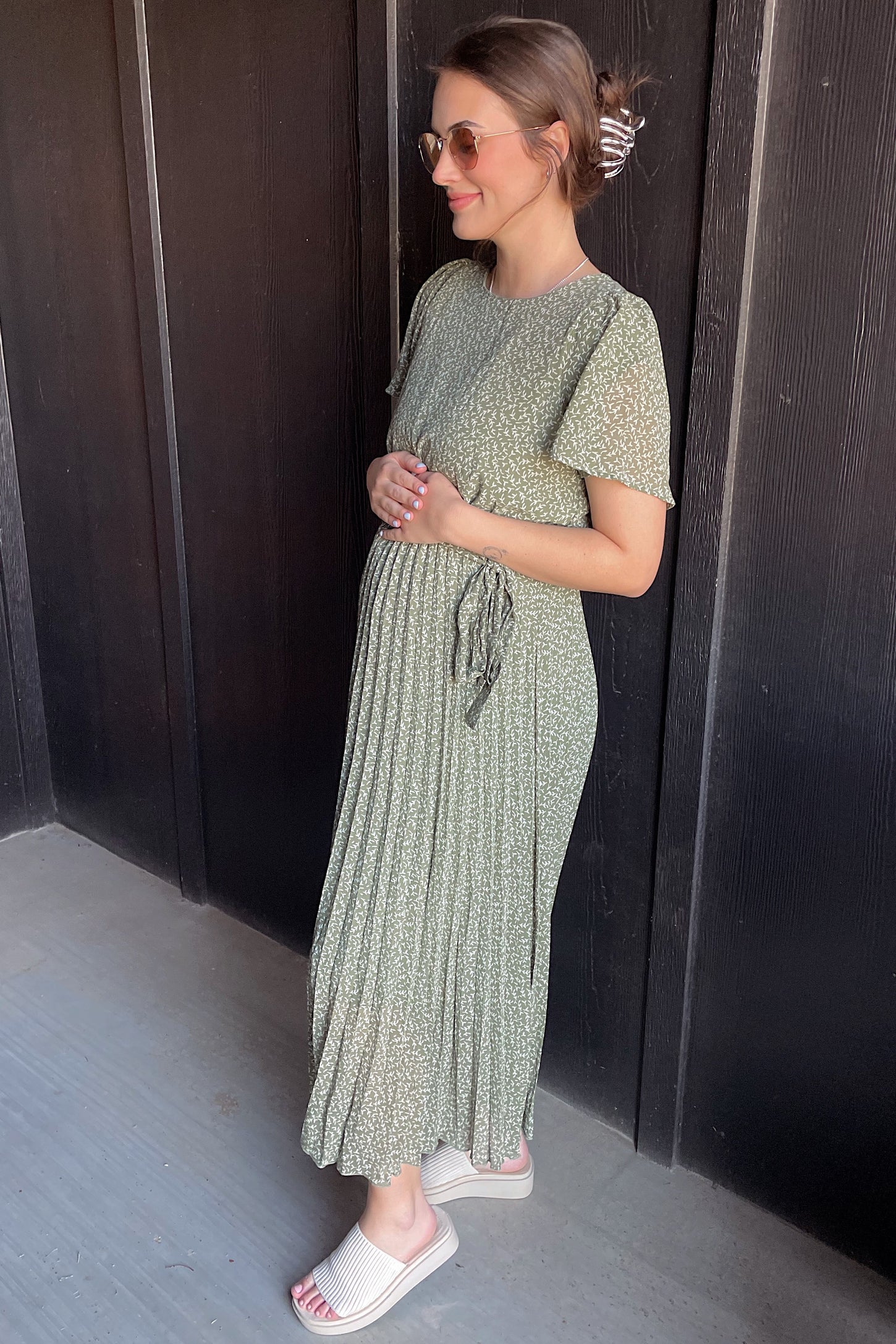 woman wearing a green floral maternity maxi dress with pleats and a tie waist, holding her baby bump