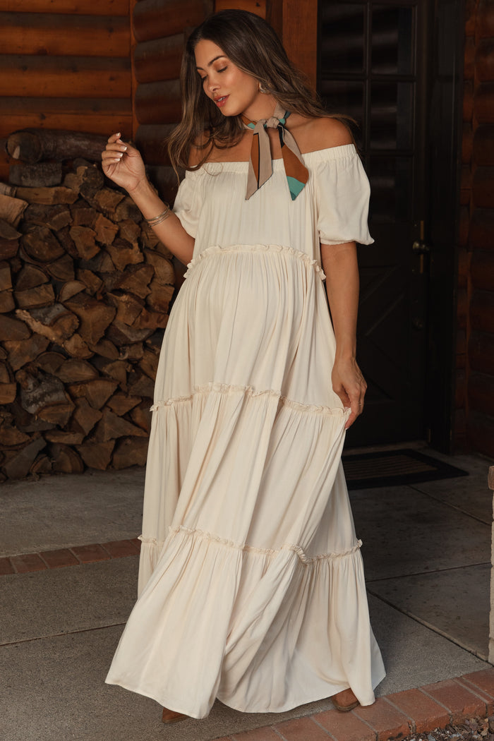 woman wearing a cream off-shoulder maternity maxi dress with tiered ruffles and a decorative neck scarf