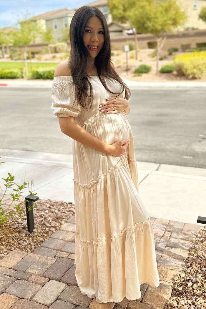woman wearing a light beige off-the-shoulder maternity dress and holding her baby bump