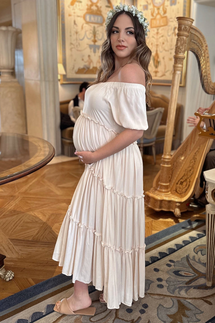 woman wearing a soft beige off-the-shoulder maternity dress with tiered details and holding her baby bump