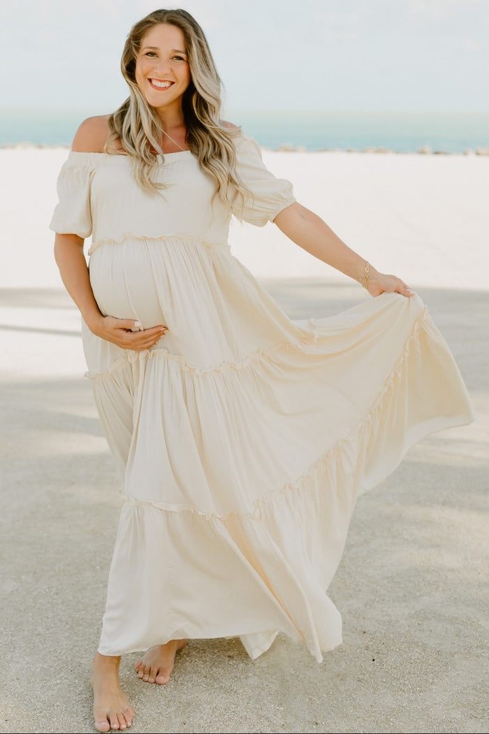 Smiling pregnant woman in a cream off-the-shoulder maternity maxi dress posing on a sandy beach