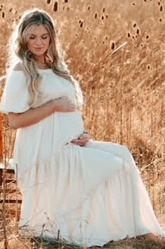 woman wearing a white off-the-shoulder maternity dress sitting in a field, holding her baby bump