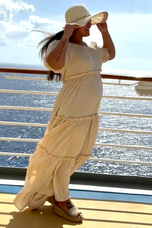 woman wearing a light beige maternity maxi dress with tiered ruffles and a wide-brimmed hat, showing her baby bump