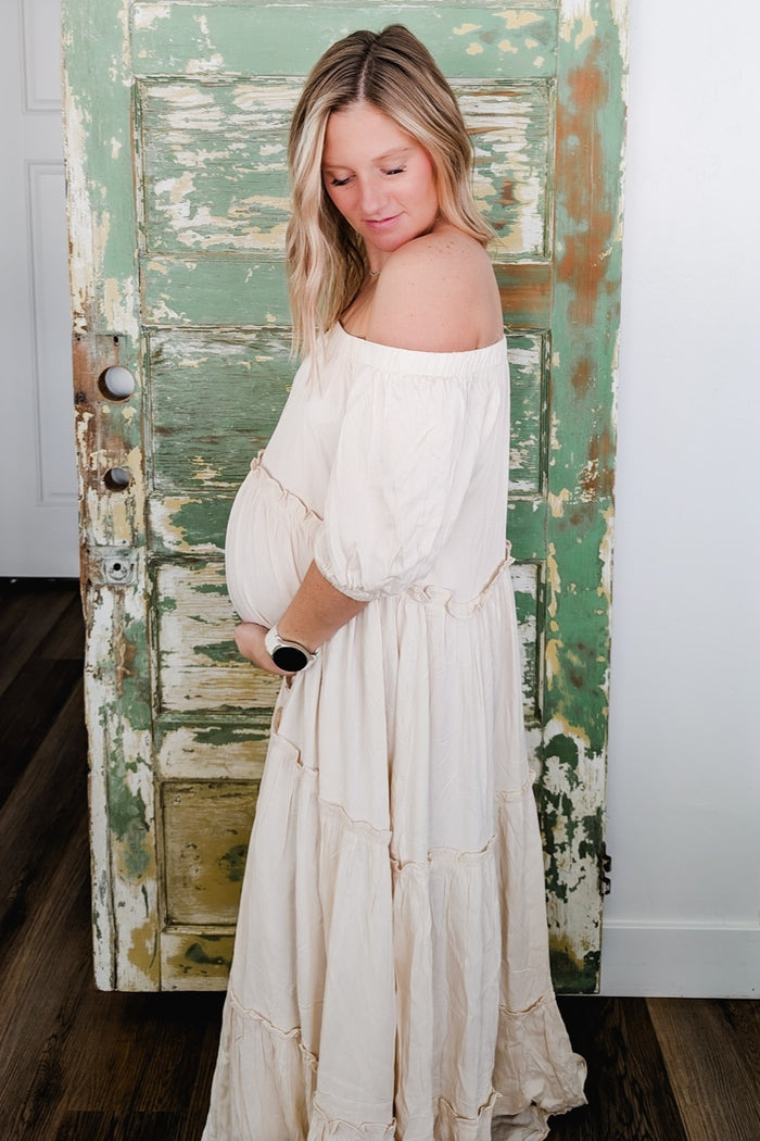 woman wearing a cream off-the-shoulder maternity dress with a flowing tiered design and holding her baby bump