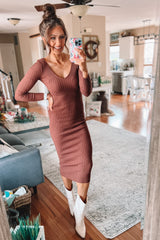 woman wearing a rust ribbed long sleeve dress and white knee-high boots, looking at the camera while posing in a stylish living room