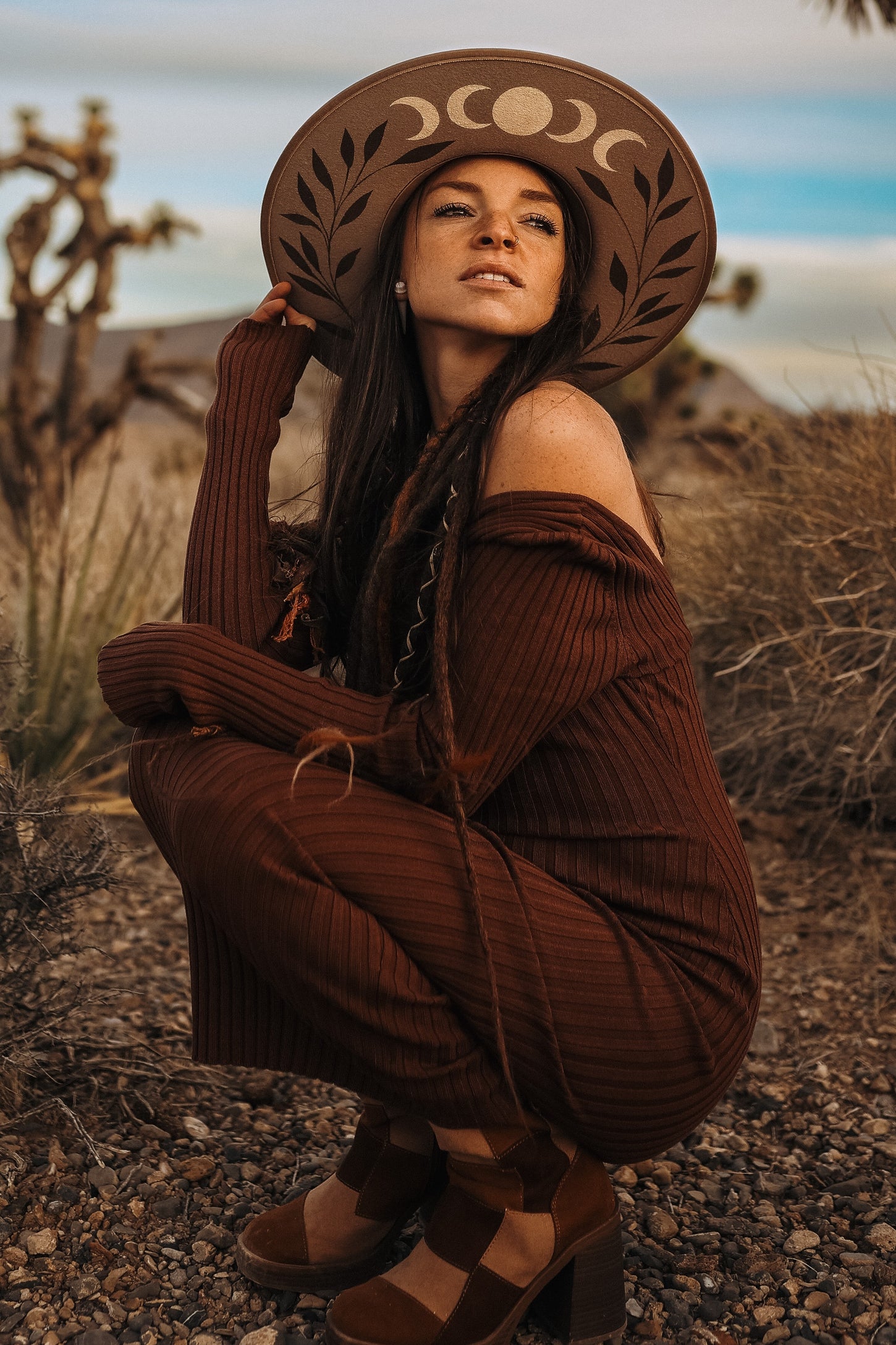 woman wearing a rust-colored off-the-shoulder ribbed dress and a wide-brimmed hat, crouching in a desert landscape