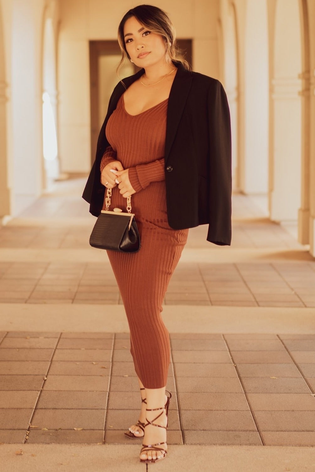 woman wearing a rust ribbed knit dress with a blazer and strappy heels, holding a black crossbody bag
