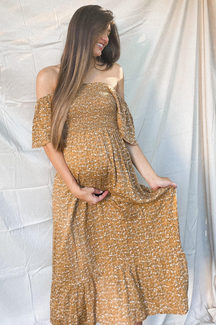 Pregnant woman in a mustard yellow floral off-the-shoulder maternity maxi dress holding her baby bump