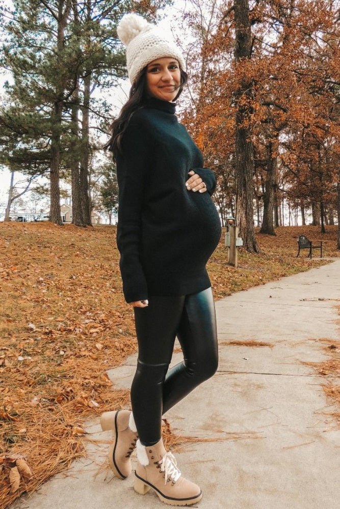 woman wearing a black maternity sweater and black leggings, holding her baby bump, walking in a park with autumn leaves.