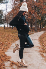woman wearing a black oversized sweater and faux leather leggings, showing her baby bump while walking on a wooded path