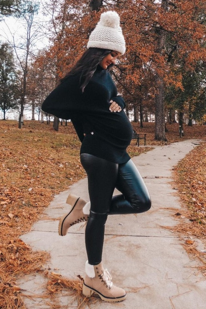 woman wearing a black oversized sweater and faux leather leggings, showing her baby bump while walking on a wooded path