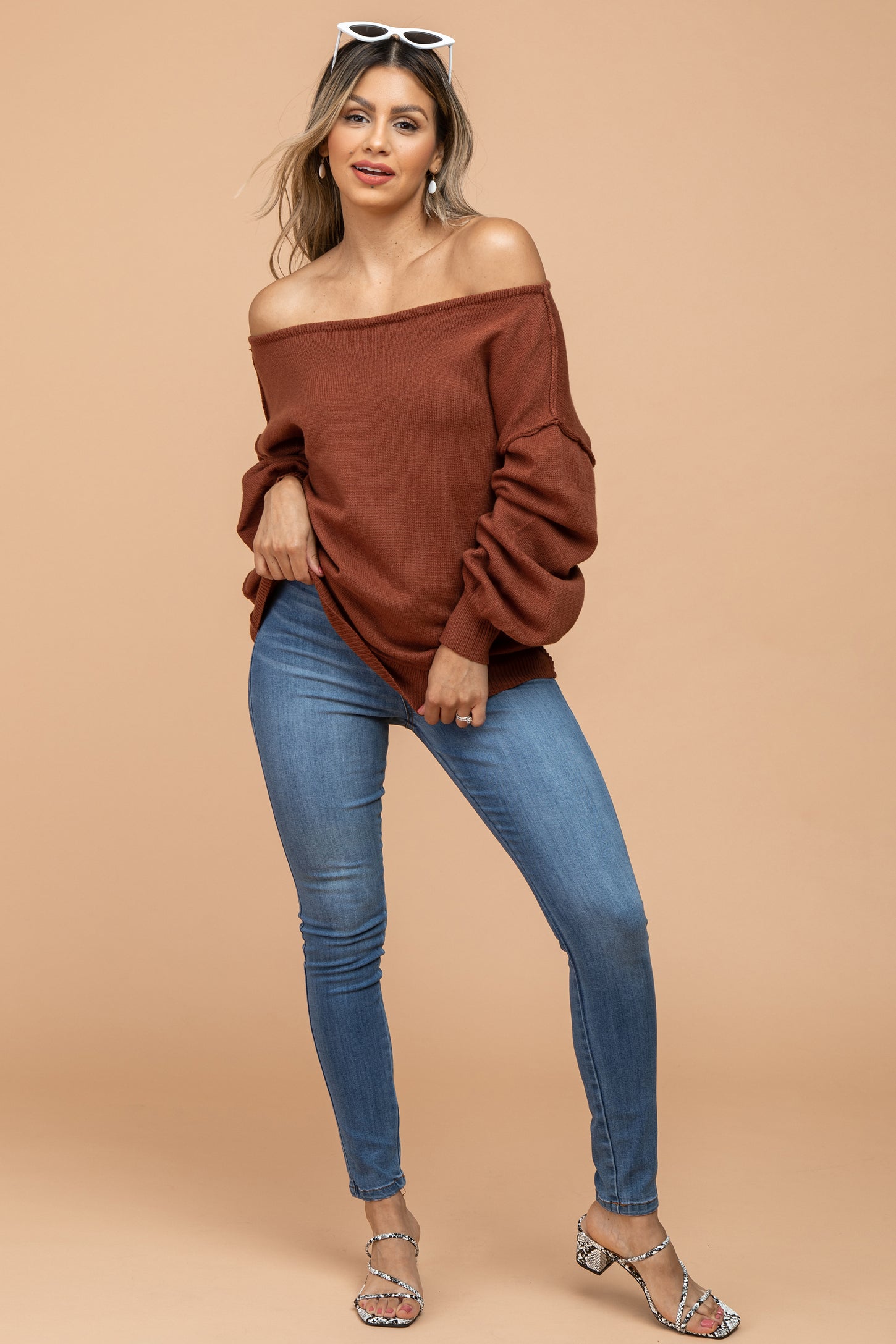 woman wearing a rust off-the-shoulder sweater and light blue skinny jeans, posing with her hand on her hip