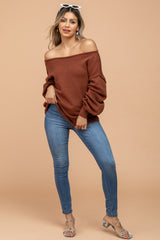 woman wearing a rust off-the-shoulder sweater and light blue skinny jeans, posing with her hand on her hip