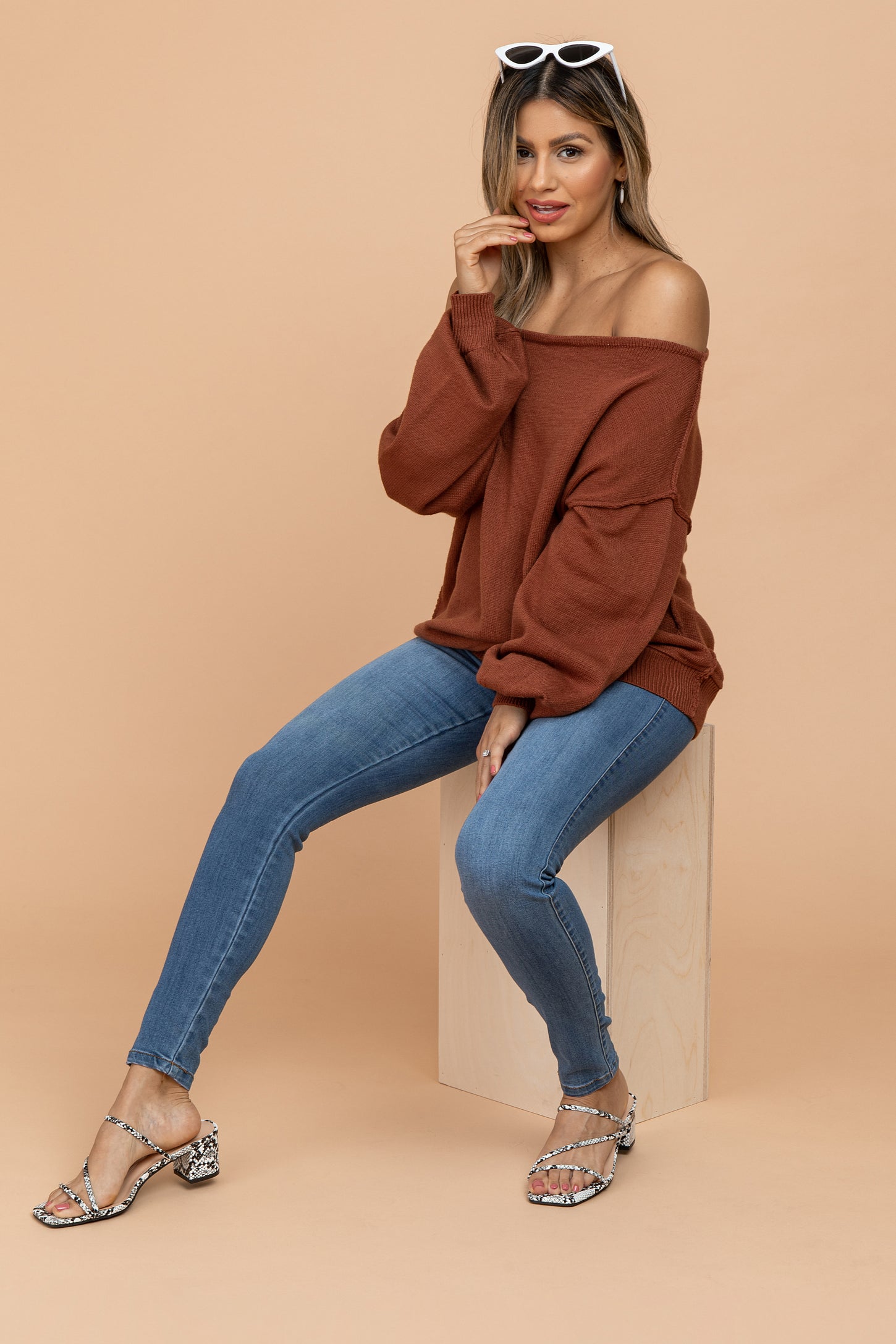 woman wearing an oversized rust off-the-shoulder sweater, blue skinny jeans, and heeled sandals, sitting on a stool