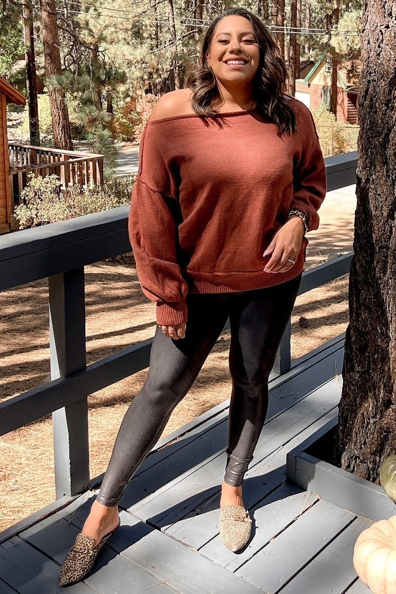 woman in rust off-the-shoulder sweater and black leggings posing on a wooden deck in a forest setting