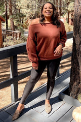woman in rust off-the-shoulder sweater and black leggings posing on a wooden deck in a forest setting