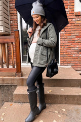 woman wearing a green maternity jacket with a baby bump, black leggings, and gray rain boots, holding an umbrella